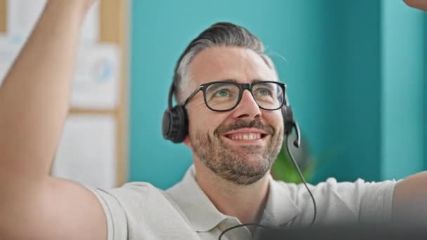 オフィスで祝うヘッドフォンを身に着けている灰色の髪の男のビジネスワーカー - 映像、動画