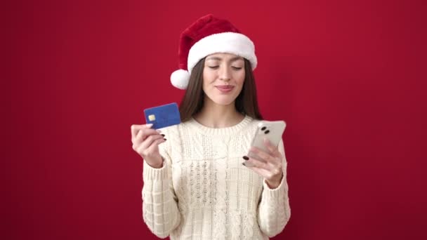 Giovane bella donna ispanica utilizzando smartphone e carta di credito in piedi su sfondo rosso isolato - Filmati, video