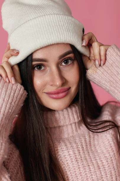 portrait of happy playful woman keeps hands on white knitted hat on pastel pink background. close-up - Fotografie, Obrázek