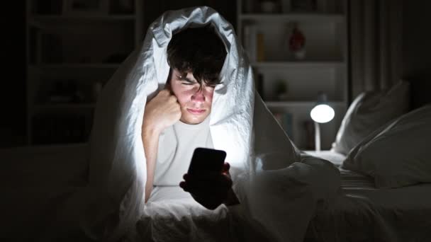 Jonge Spaanse tiener serieus bezig met sms 'en op zijn smartphone, liggend op een comfortabel bed onder een knusse deken, in het schemerige licht van zijn gezellige slaapkamer - Video