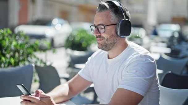 Седовласый мужчина, слушающий музыку, сидящую на столе на террасе кафе - Кадры, видео
