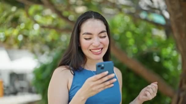 Junge schöne hispanische Frau feiert mit Smartphone im Park - Filmmaterial, Video