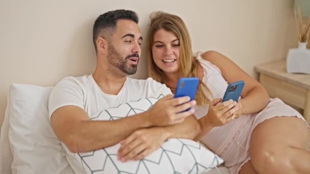 Pareja de hombres y mujeres sentados en la cama usando teléfonos inteligentes hablando en el dormitorio - Imágenes, Vídeo