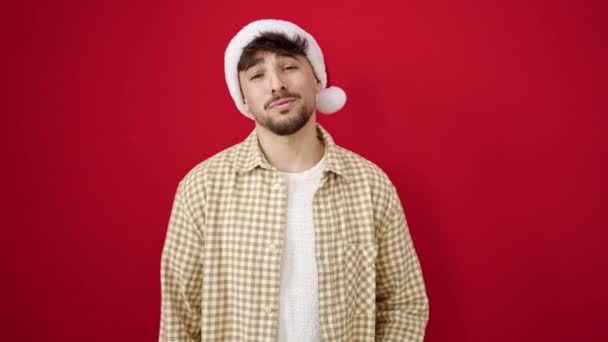 孤立した赤い背景の上にクリスマスの帽子を身に着けている自信を持って微笑む若いアラブの男 - 映像、動画