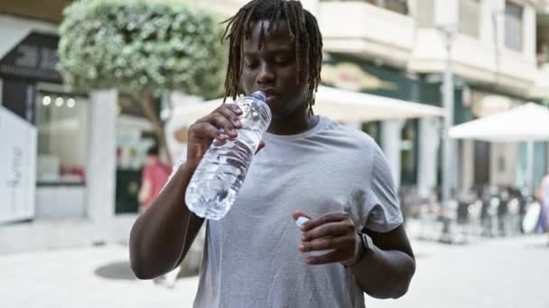 Uomo afroamericano che beve bottiglia d'acqua facendo gesto pollice su alla terrazza della caffetteria - Filmati, video