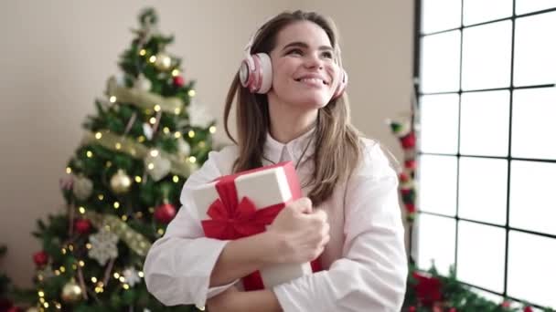 Νεαρή όμορφη ισπανόφωνη γυναίκα χορεύει στο χριστουγεννιάτικο δέντρο κρατώντας δώρο στο σπίτι - Πλάνα, βίντεο
