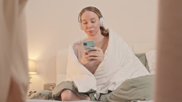 Νεαρή λευκή γυναίκα κάθεται στο κρεβάτι με κουβέρτα στους ώμους της, απολαμβάνοντας μουσική σε ασύρματα ακουστικά και χρησιμοποιώντας smartphone, περνώντας το πρωί στο σπίτι μόνη της - Πλάνα, βίντεο