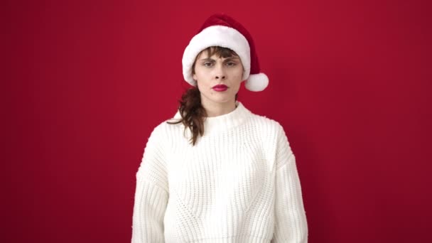 Mujer caucásica joven haciendo signo negativo con los pulgares hacia abajo usando sombrero de Navidad sobre fondo rojo aislado - Imágenes, Vídeo