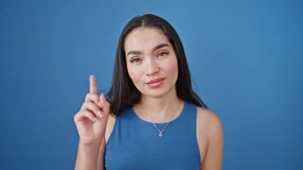 Νεαρή όμορφη ισπανόφωνη γυναίκα χαμογελά με αυτοπεποίθηση λέγοντας όχι με το δάχτυλο πάνω από απομονωμένο μπλε φόντο - Πλάνα, βίντεο