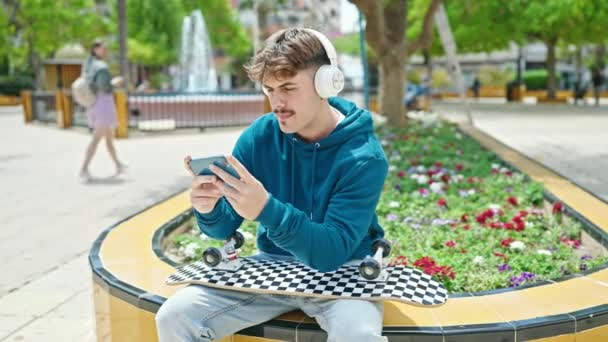 Νεαρός Ισπανός που κρατάει πατινάζ παίζοντας βιντεοπαιχνίδι στο πάρκο - Πλάνα, βίντεο