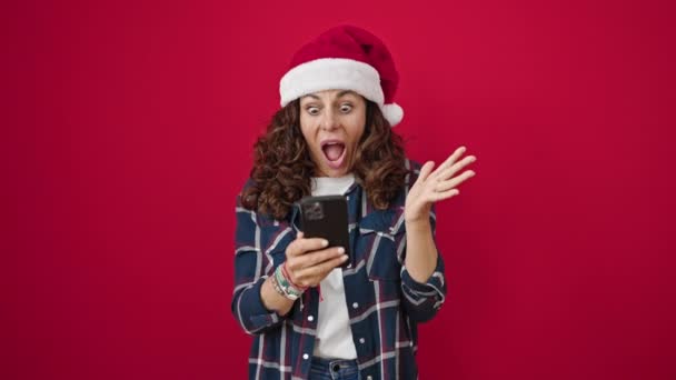 Noel şapkası takan orta yaşlı İspanyol kadın izole edilmiş kırmızı arka planda akıllı telefona işaret ediyor. - Video, Çekim