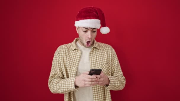 Молодой латиноамериканец в рождественской шляпе указывает на смартфон на изолированном красном фоне - Кадры, видео
