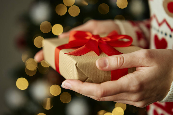 Egy meleg pulóveres nő, aki ajándékdobozt tart kézműves papírba csomagolva ünnepi szalaggal. Ajándék a téli szünetre. Karácsony és boldog új évet ünneplő - Fotó, kép