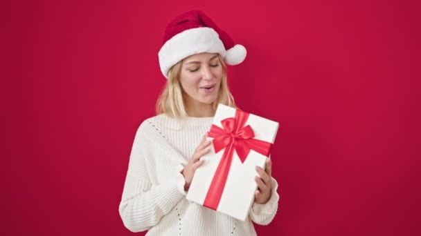 fiatal szőke nő visel karácsonyi kalap gazdaság ajándék felett elszigetelt piros háttér - Felvétel, videó
