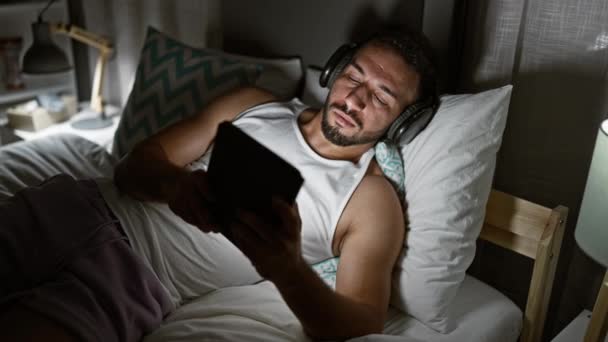 Hombre árabe joven usando touchpad acostado en la cama con auriculares en el dormitorio - Imágenes, Vídeo