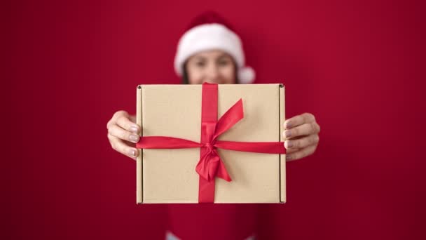 Genç, güzel, İspanyol bir kadın Noel şapkası takıyor ve kırmızı arka planda hediyeler tutuyor. - Video, Çekim