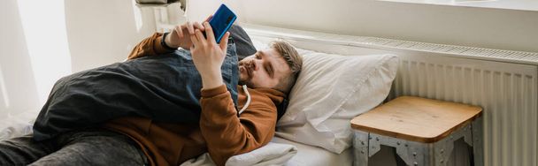 Фрилансер, пользующийся мобильным телефоном, проверяет каналы социальных сетей или чаты сообщений, сидя дома на кровати. Цифровое онлайн-общение отдыхает после тяжелого рабочего дня - Фото, изображение