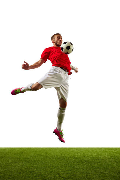 Genç bir adam, kırmızı spor giyimli profesyonel bir futbolcu ve yeşil çimenli beyaz arka plana karşı futbol numaraları yapıyor. Oyun, spor, eğlence, aktif yaşam tarzı kavramı. - Fotoğraf, Görsel