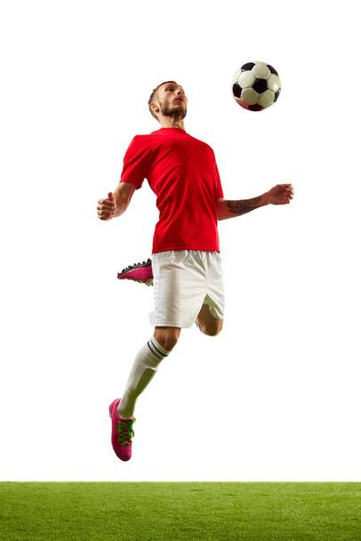 Young man, professional player in red sportwear and boots training football tricks against white background with green grass. Concept of game, sport, recreation, active lifestyle. - Photo, Image