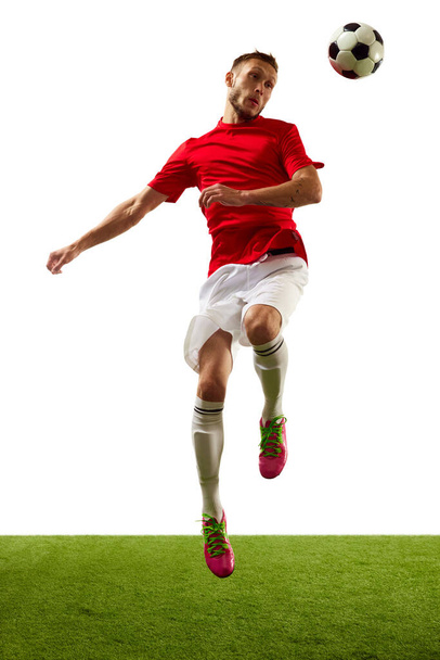 Енергійний молодий чоловік, професійний гравець у червоному спортивному одязі та чоботях, тренує футбольні трюки на білому тлі з зеленою травою. Концепція гри, спорту, відпочинку, активного способу життя. - Фото, зображення
