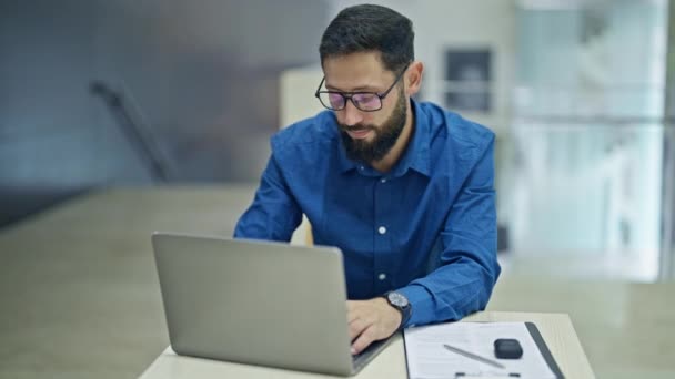 Νεαρός ισπανόφωνος επιχειρηματίας που χρησιμοποιεί φορητό υπολογιστή που βγάζει γυαλιά στο γραφείο - Πλάνα, βίντεο
