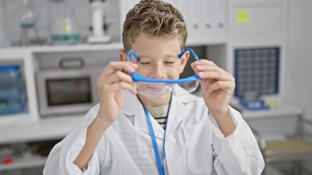 Adorable blond boy, confidently smiling working indoors as a scientist, wearing lab glasses in the center of a bustling laboratory, showcasing the joy of childhood medicine research. - Footage, Video
