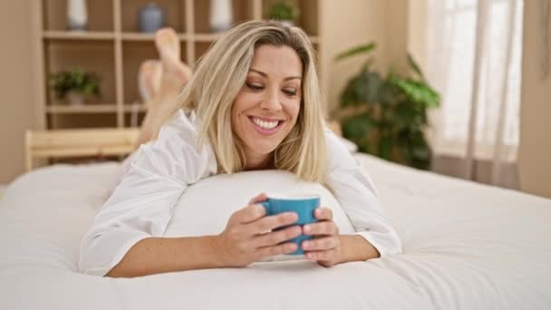 Nuori blondi nainen juo kuppi kahvia makaa sängyssä makuuhuoneessa - Materiaali, video