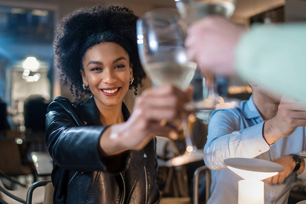 Promienna młoda kobieta z fryzurą Afro radośnie wznosi toast kieliszkiem do wina w nowoczesnym barze, uchwycając uroczysty i elegancki moment. - Zdjęcie, obraz