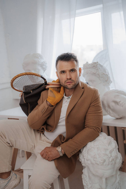Stylish male model embodies the old-money look, posing with a bag and tennis racket among classical Greek sculptures in a sculptors atelier - Photo, Image