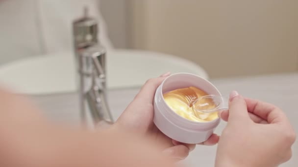 Donna irriconoscibile che prende idrogel d'oro sotto le toppe degli occhi dal vaso con cucchiaio di plastica mentre fa la routine di cura della pelle in bagno - Filmati, video