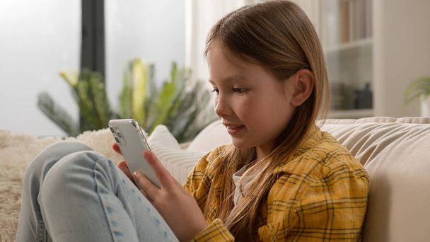 Χαμογελώντας χαρούμενο κορίτσι παιδί στέλνοντας μήνυμα ελέγχοντας email χρησιμοποιώντας κινητό τηλέφωνο χαριτωμένο σχολιαρόπαιδο chatting με τους φίλους στο κοινωνικό δίκτυο internet παίζοντας online παιχνίδι βλέποντας βίντεο στο smartphone στο σπίτι - Φωτογραφία, εικόνα