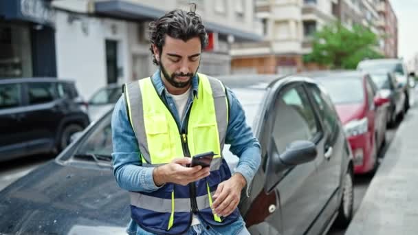 Joven hispano de pie en coche con chaleco reflectante usando smartphone en la calle - Imágenes, Vídeo