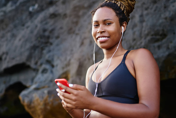 Портрет позитивної спортивної жінки, яка посміхається і слухає мотиваційний плейлист перед початком ранкового тренування, підключеного до 4g бездротового зв'язку на мобільному телефоні для використання в Інтернеті
 - Фото, зображення