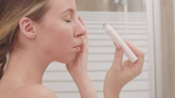 Medium closeup of pretty young natural Caucasian woman applying eye roller serum while standing in front of mirror in bathroom wearing white towel on her body - Footage, Video