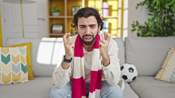 Young hispanic man watching soccer match crossing fingers celebrating at home - Footage, Video
