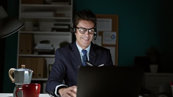 Joven trabajador de negocios hispano usando laptop y auriculares celebrando en la oficina - Metraje, vídeo