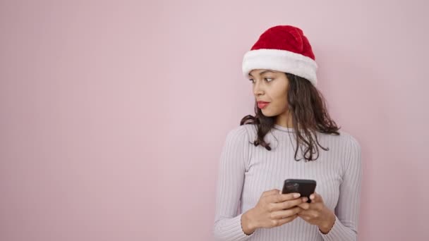 Νεαρή όμορφη ισπανόφωνη γυναίκα φορώντας χριστουγεννιάτικο καπέλο χρησιμοποιώντας smartphone πάνω από απομονωμένο ροζ φόντο - Πλάνα, βίντεο