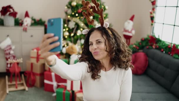 Hispanische Frau mittleren Alters macht Selfie mit dem Smartphone, wenn sie zu Hause am Weihnachtsbaum auf dem Boden sitzt - Filmmaterial, Video