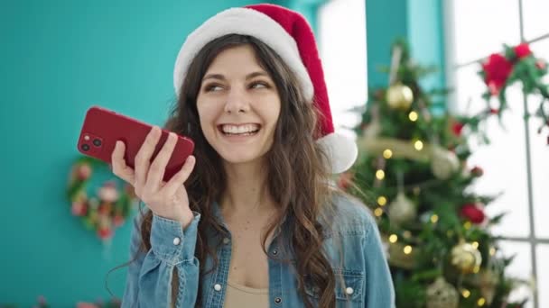 Молодая красивая латиноамериканка отправляет голосовое сообщение на смартфоне, празднуя Рождество дома - Кадры, видео