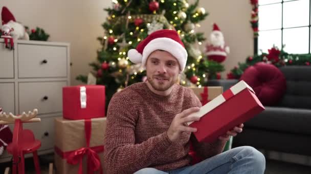 Молодий білий чоловік тримає подарунок, сидячи на ялинці вдома - Кадри, відео
