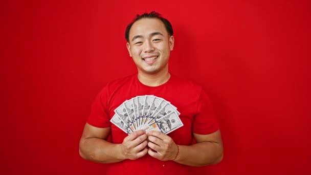 Homem chinês jovem confiante, brilhando de felicidade, casualmente segurando um monte de nós dólares, sorrindo corajosamente sobre um fundo vermelho isolado. - Filmagem, Vídeo