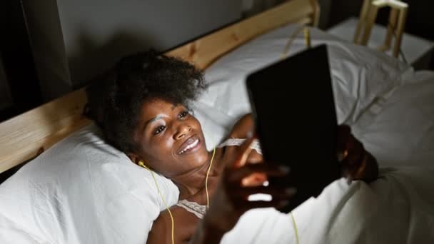 Afrikai amerikai nő használ touchpad és fülhallgató feküdt az ágyon mosolyogva a hálószobában - Felvétel, videó