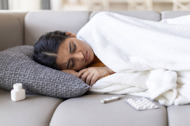 Mujer india joven enferma que duerme con fiebre alta en el sofá en casa, mujer oriental enferma acostada debajo de una manta junto a las píldoras de la medicina, sintiéndose mal, sufriendo gripe estacional y frío, disparo de primer plano - Foto, Imagen