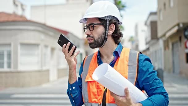 Mladý hispánský muž architekt držící plány posílající hlasovou zprávu chytrým telefonem na staveništi - Záběry, video
