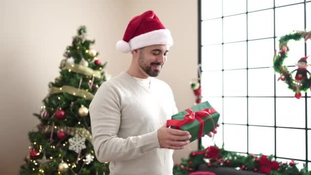 Junger hispanischer Mann umarmt Geschenk, das zu Hause am Weihnachtsbaum steht - Filmmaterial, Video