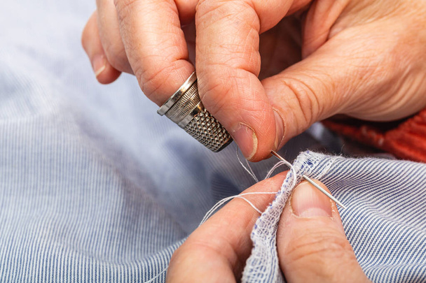 Close up photo extreme horizontal, hands mature adult Caucasian woman. sewing with needle and stitch, to repair a blue shirt, doing tailor's manual work. - Photo, Image