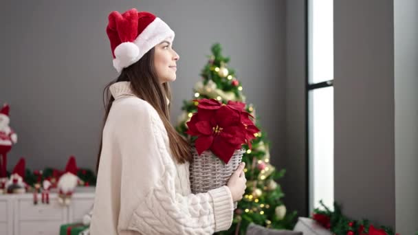Νεαρή όμορφη ισπανόφωνη γυναίκα κρατώντας το φυτό στέκεται δίπλα στο χριστουγεννιάτικο δέντρο στο σπίτι - Πλάνα, βίντεο