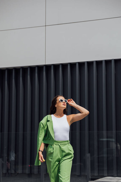 Módní elegantní sebevědomá žena v módních růžových slunečních brýlích, hedvábném šátku, sako, se zelenou taškou, pózující na ulici evropského města. Venkovní módní portrét. Kopírovat, prázdné místo pro text - Fotografie, Obrázek
