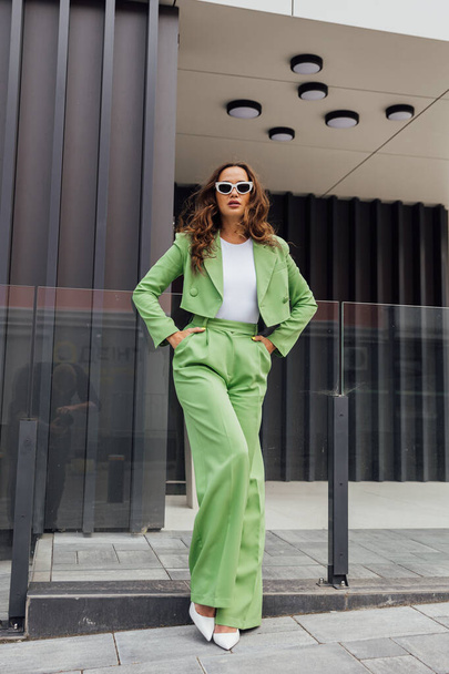 Fashionable elegant confident woman wearing trendy pink sunglasses, silk headscarf, suit blazer, with green bag, posing in street of European city. Outdoor fashion portrait. Copy, empty space for text - Foto, Imagen