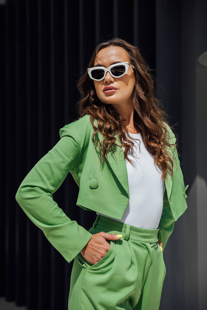 Fashionable elegant confident woman wearing trendy pink sunglasses, silk headscarf, suit blazer, with green bag, posing in street of European city. Outdoor fashion portrait. Copy, empty space for text - Photo, Image